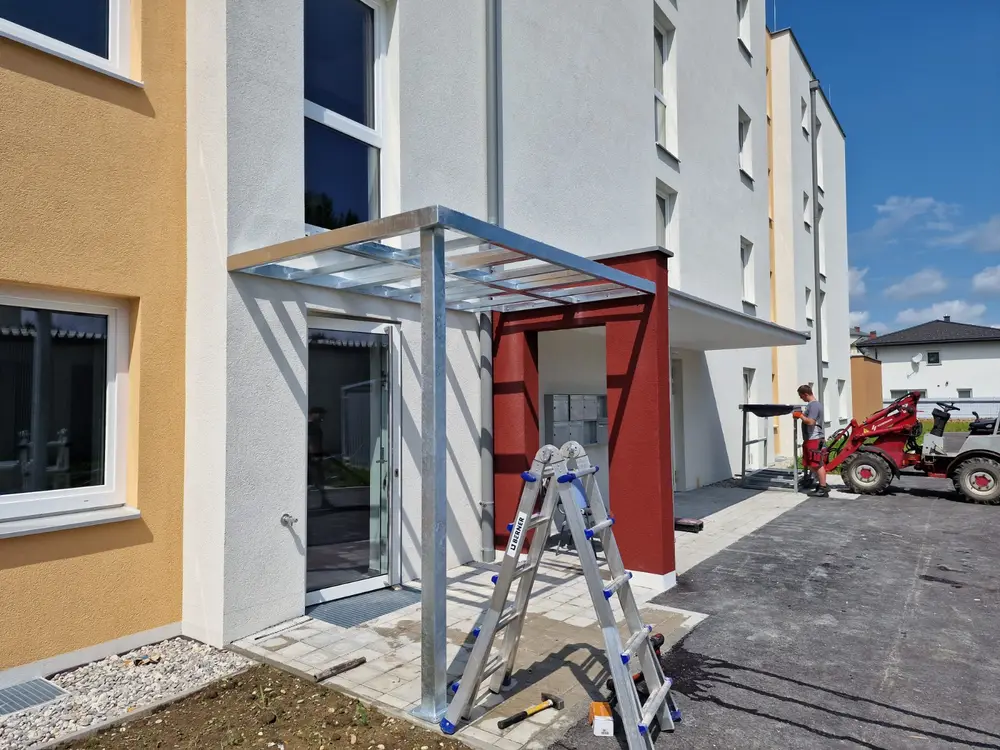 Vordach aus Stahl und Glas über dem Hauseingang eines Wohnblocks, Leiter und Hoflader auf der Baustelle