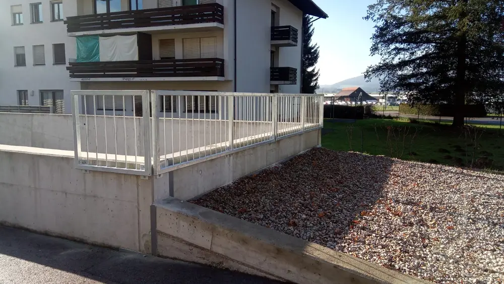 Weißes Terrassengeländer mit senkrechten Stäben auf einer Betonbrüstung, Wohnhaus im Hintergrund