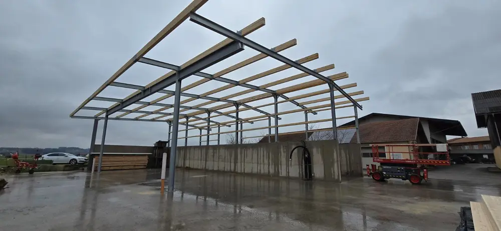 Stahlrahmen eines Unterstands mit aufgelegten Holzpfetten, Betonwand und Hofplatz im Regen